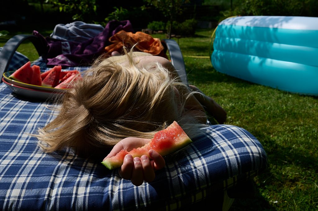 Junge mit längeren Haaren liegt auf einer Liege in der Sonne. Er hält eine Stück abgeknabberte Wassermelone in der Hand. Im Hintergrund steht ein Pool.